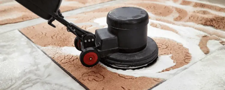 Close-up of a heavy-duty rotary floor scrubber machine applying a thick, eco-friendly foam to deep clean a textured rug. The image illustrates professional agitation techniques used to lift dirt and allergens from carpets and area rugs.
